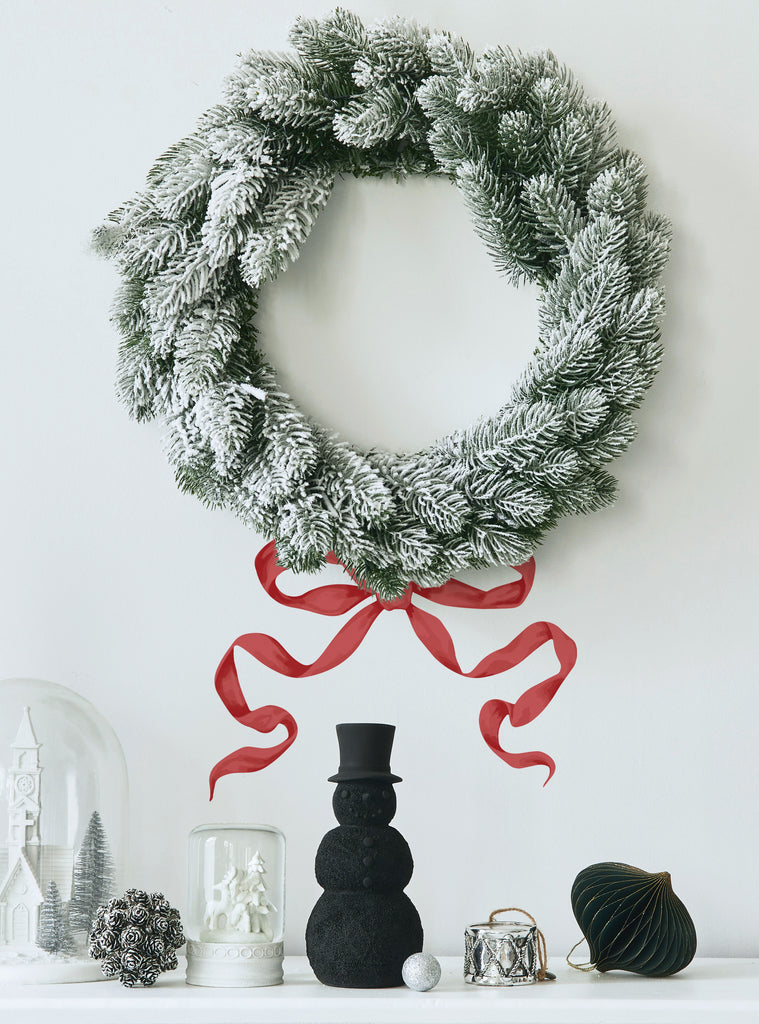 Snowy green holiday wreath hanging on wall with festive red ribbons stickers and winter decor below on shelf