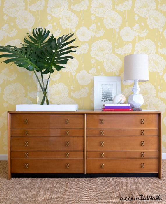 Modern yellow peony wallpaper with detailed blooms behind a wooden dresser styled with a lamp, books, and tropical leaves in a vase