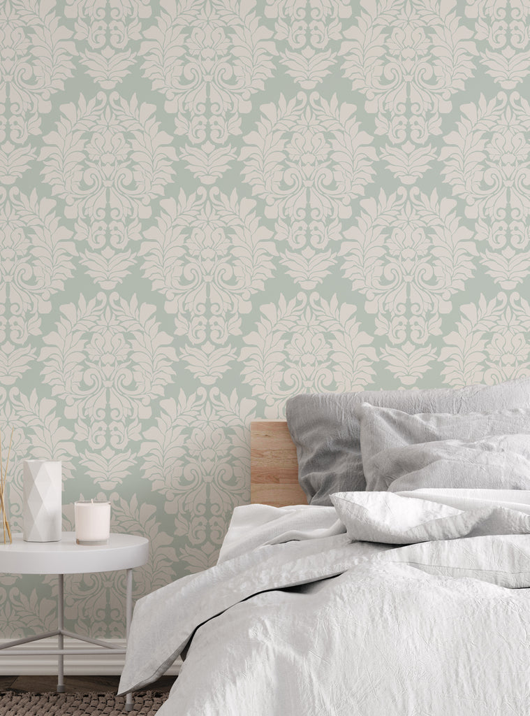 Light green and beige damask classic peel and stick wallpaper shown in a cozy bedroom setting with soft bedding and a side table.