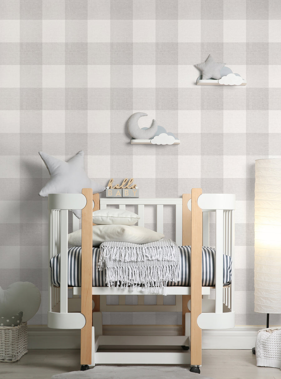 The image shows a cozy and modern nursery setting with a checkered fabric wallpaper in a neutral color palette. The room features a white crib with soft textiles and accessories, including plush toys resembling clouds. This image would be suitable for a Shopify product page showcasing the "Checkered Fabric Look Medium Peel and Stick Wallpaper" by highlighting its versatile and homey aesthetic, perfect for creating a warm and inviting space. This 
