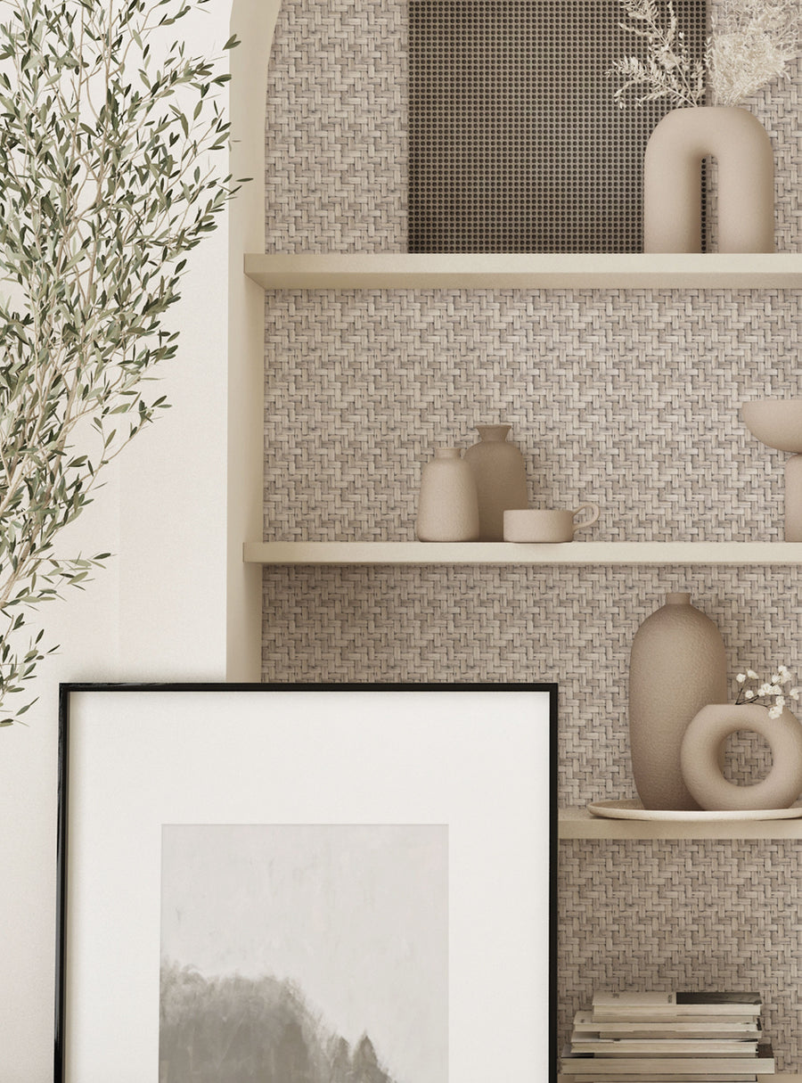 A cozy interior scene featuring a light-colored woven wall as a backdrop for minimalist decor. Shelves display various ceramic vases and a circular object, alongside a framed artwork with muted tones. A leafy plant is partially visible on the left side, adding a touch of greenery.