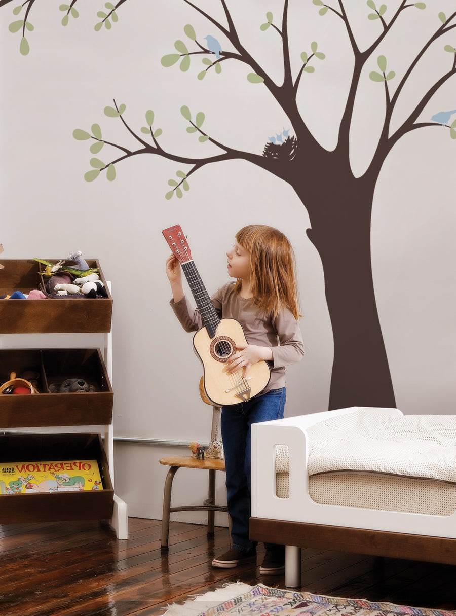 A young girl playing a guitar in a room with a large tree mural on the wall, featuring birds and a nest, creating a whimsical and nature-inspired atmosphere. , Featured Collection,Top Sellers,Wall Decals,Animal Decals,Baby Nursery and Kids Wall Decals,All Products