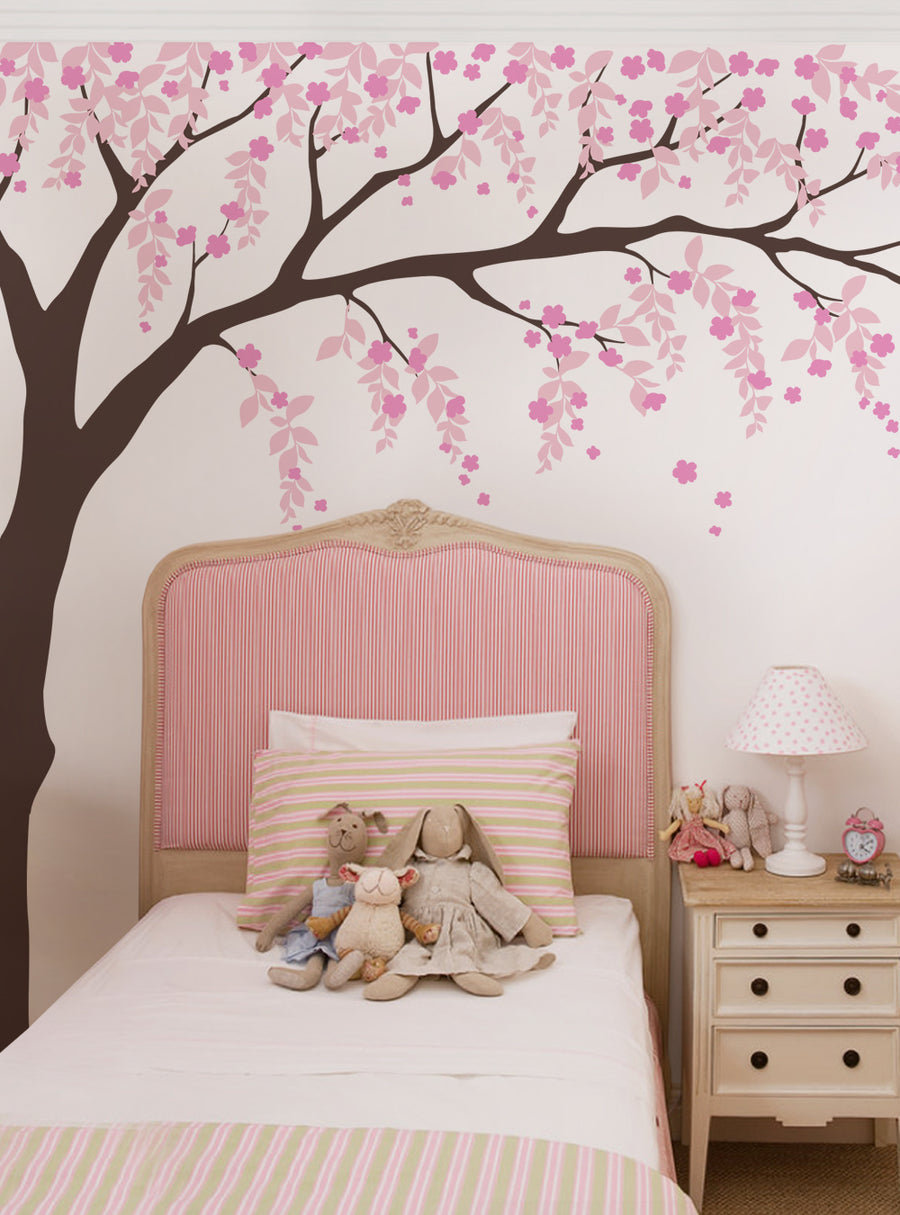 Alt text: A mural depicting a weeping willow tree with cascading cherry blossoms adorns the wall behind a pink-striped headboard in a cozy, feminine bedroom setting. The tree's branches and flowers create a soothing, nature-inspired atmosphere. This wall decal would add elegance and charm to any living space. Top Sellers,Wall Decals,Floral Decals,Cherry Blossom Tree Decals,Tree Decals,Baby Nursery and Kids Wall Decals,All Products