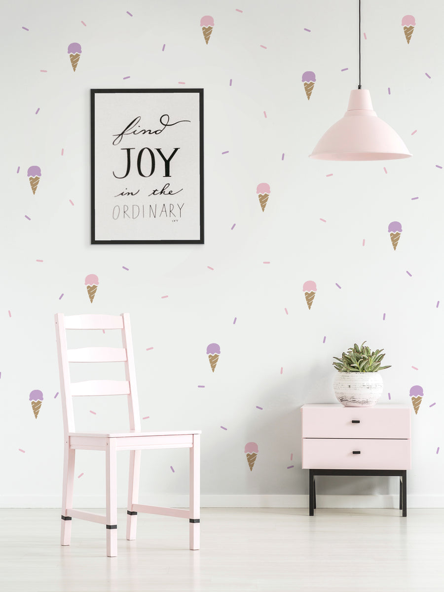 Alt text: This image depicts a minimalist interior design with a wall featuring an inspirational quote framed in black, "Find Joy in the Ordinary", along with a repeating pattern of colorful ice cream cone illustrations in various shades of pink, purple, and blue. The room also includes a pale pink chair, a pink and black dresser, and a hanging light fixture. This wall decal would be a charming addition to a baby nursery, kid's room, or any space