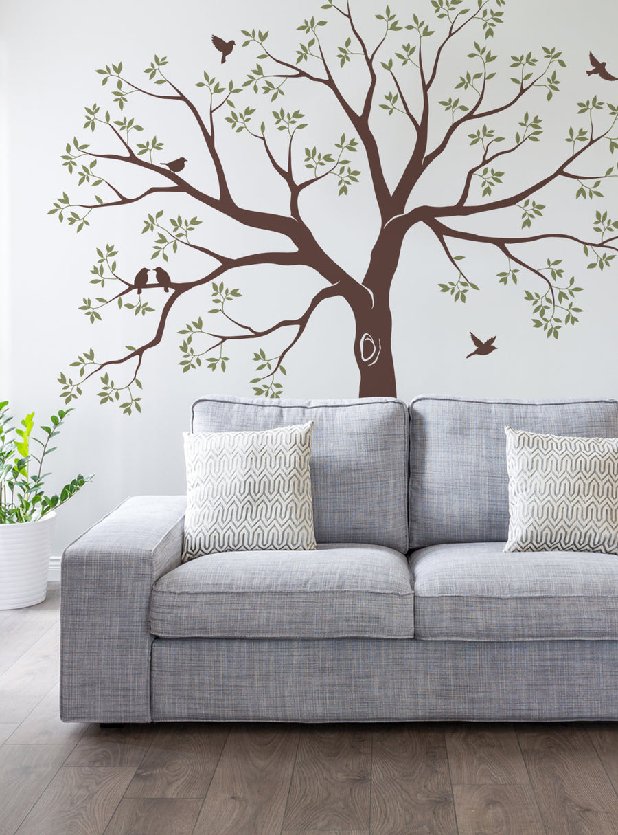 This detailed image depicts a large tree wall decal featuring a silhouetted tree with numerous branches and leaves, as well as several birds perched on or flying around the tree. The tree appears to be a focal point of the room's decor, complementing the neutral-toned sofa and surroundings. This staircase family tree wall decal would be an eye-catching and memorable addition to any living space, providing a natural, rustic, and sentimental touch 