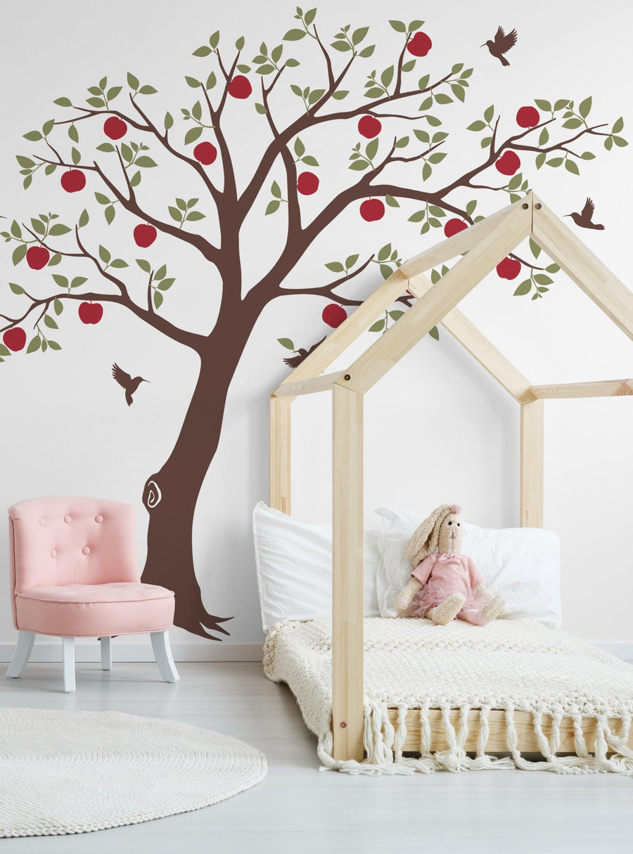 Elaborative Alt Text: A vibrant fruit tree with lush green leaves and ripe red apples serves as a stunning focal point in this cozy and inviting nursery space. The tree's branches reach upwards, creating a whimsical and charming backdrop for the adorable bed frame and pink accent chair. Hummingbirds flutter amidst the foliage, adding a touch of natural wonder to the scene. This enchanting wall decal effortlessly blends nature and imagination, mak