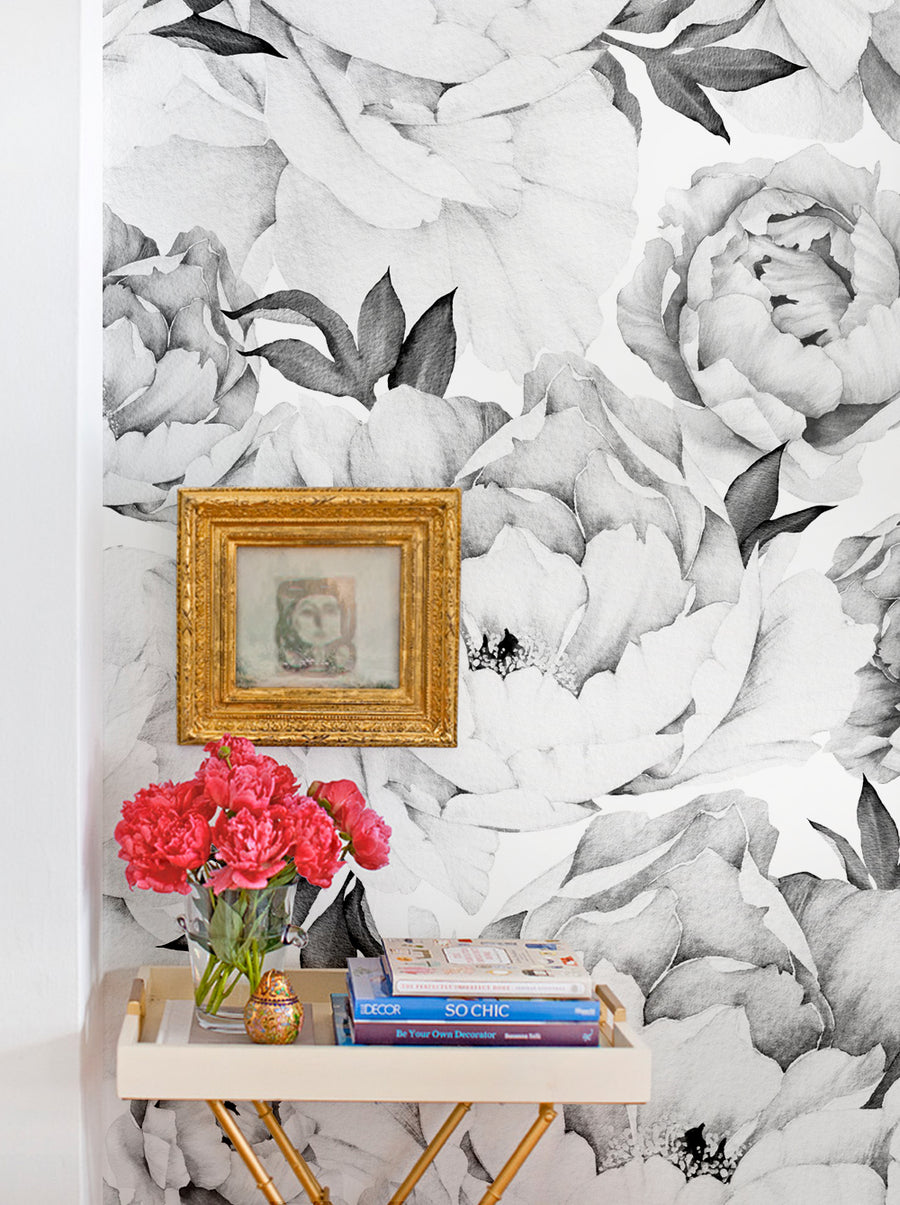 A stylish corner featuring black and white floral wallpaper with peonies. In the foreground, there's a small table holding a vase of vibrant pink flowers and a stack of books. A framed artwork in a gilded frame hangs on the wall, adding an artistic touch to the decor. The combination of the wallpaper and decorative elements creates a chic and inviting atmosphere.