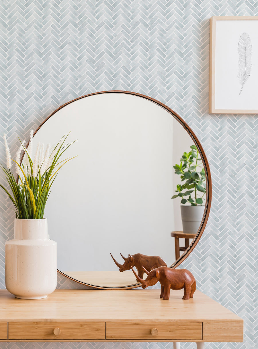 A large circular mirror with a wooden frame, set against a herringbone patterned tile wall. On the wooden console below, there are various decorative elements including a potted plant, vase, and two wooden elephant figurines. This image showcases a tastefully styled vignette that highlights the herringbone wallpaper and reflects the natural light in the room., Top Sellers,Peel and Stick Wallpaper,Geometric & Pattern Wallpaper,All Products,Unpaste