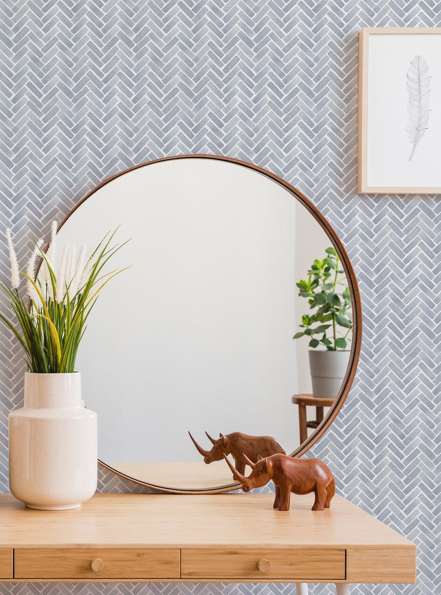 The image depicts a stylish and modern interior design setting, featuring a round mirror with a wooden frame, a wooden console table with drawers, and a variety of decorative accents such as a plant, a vase, and two small elephant figurines. The background wall has a herringbone pattern in shades of gray, creating a sophisticated and textural backdrop. This alt text describes the key elements of the image without identifying any individuals prese
