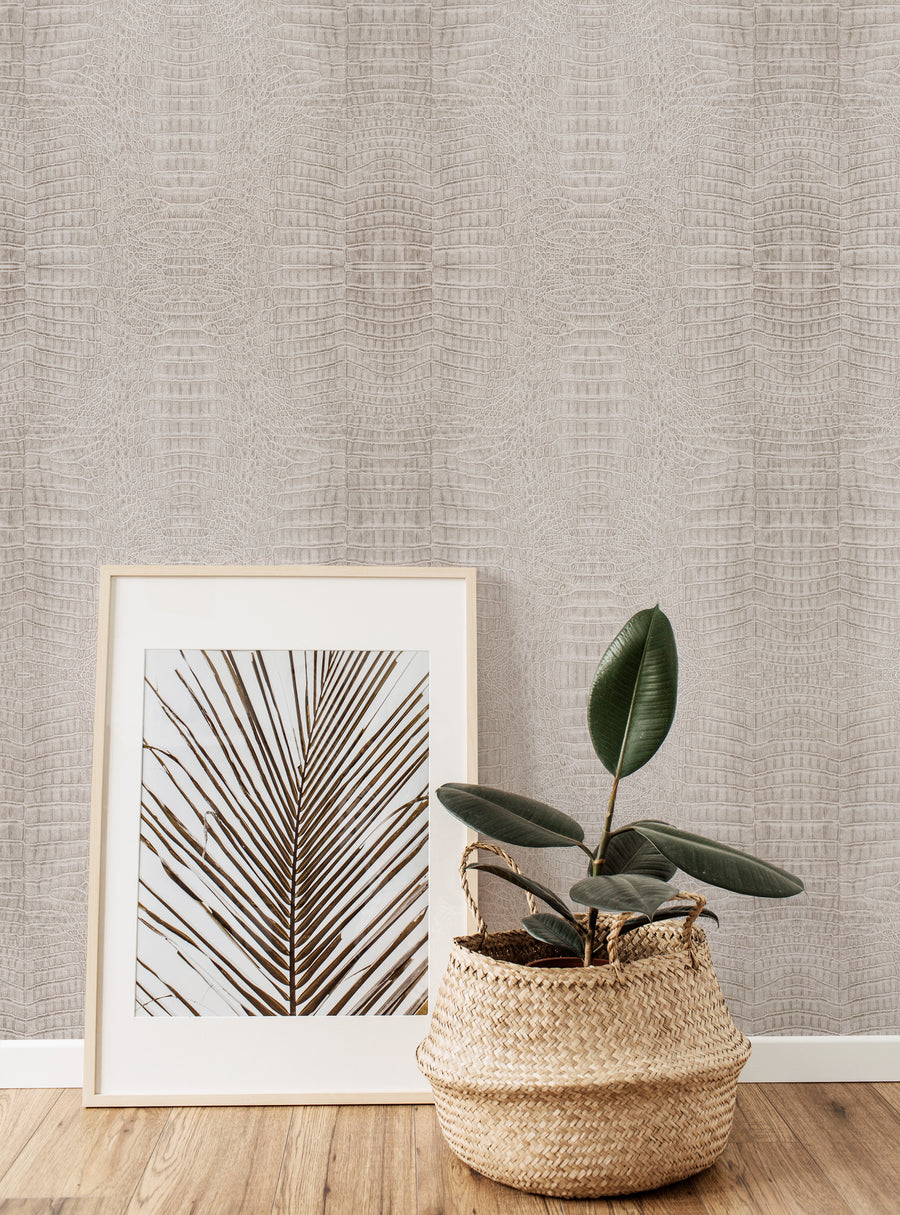 A stylish interior featuring beige crocodile-patterned wallpaper. In the foreground, a framed artwork displaying a palm leaf design is positioned against the wall. Beside it, a woven basket contains a green plant, adding a natural element to the decor. The combination of textures and neutral tones creates a warm and inviting atmosphere, enhancing the overall aesthetic.