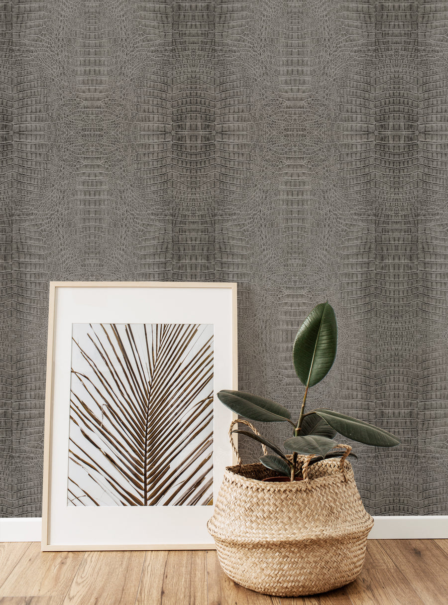 A stylish corner featuring grey crocodile-patterned wallpaper. In the foreground, a framed artwork of a palm leaf rests against the wall. Next to it, a woven basket planter holds a green plant with large, glossy leaves. The combination of natural elements and the textured wallpaper creates a harmonious and modern atmosphere. The wooden floor adds warmth to the overall aesthetic.