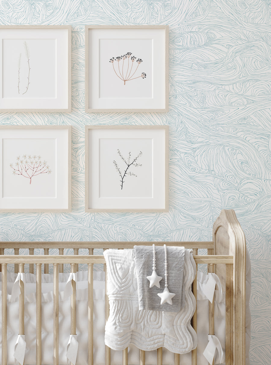 Image of abstract botanical sketches framed on a wall above a crib in a nursery. Soft, natural colors and textures create a calming, nature-inspired atmosphere. Described as "Wood Grain Waves Peel and Stick Wallpaper", the wallpaper features a hand-drawn sketch style with intricate lines and shading that mimics the look of real wood grain. This versatile and artistic wallpaper design would complement a variety of styles, from modern to rustic, in